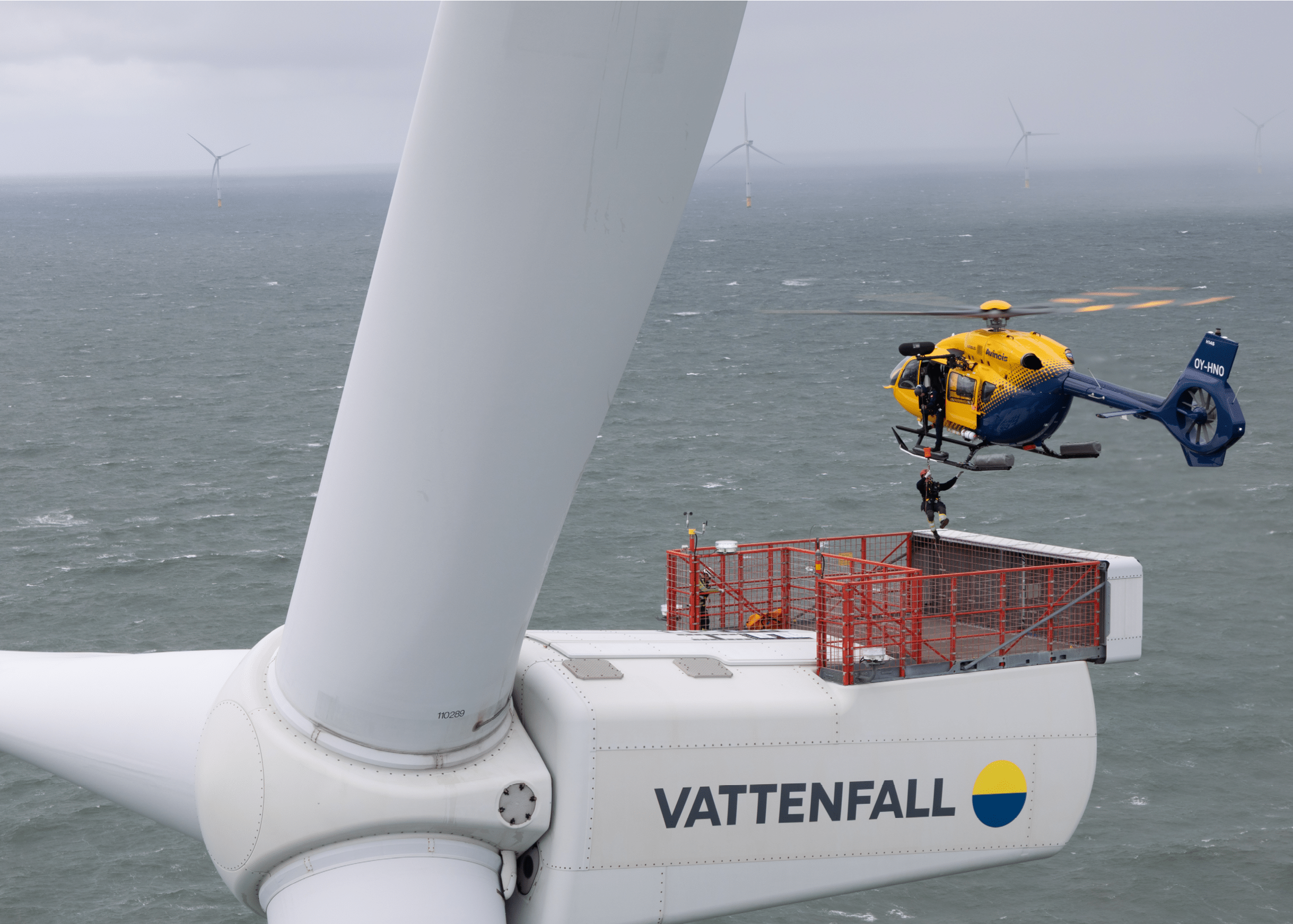 A helicopter is hovering next to the top of a large wind turbine labeled Vattenfall above a body of water with other wind turbines visible in the distance. Description generated by AI