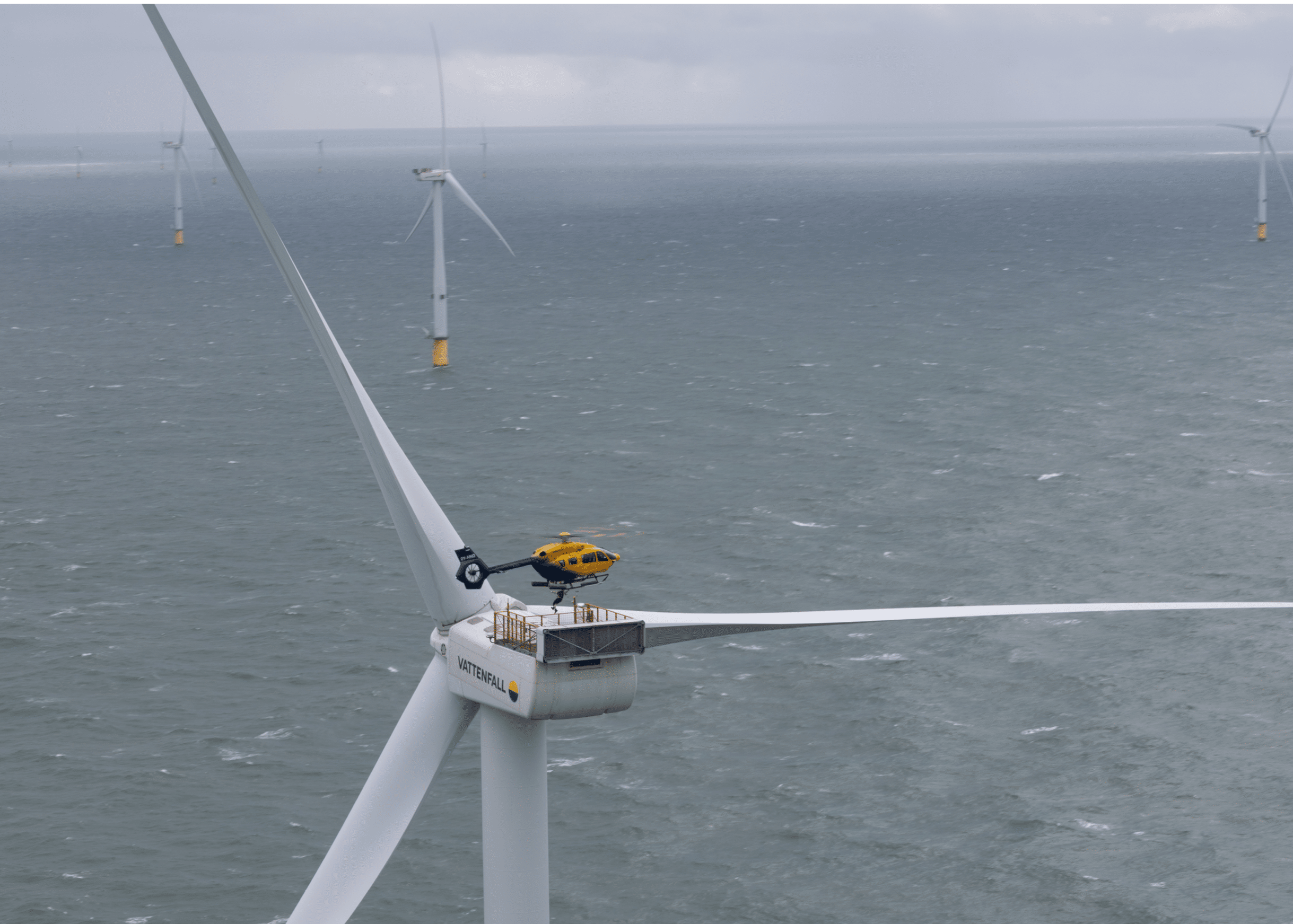 A yellow helicopter is landing on top of a large offshore wind turbine surrounded by other wind turbines in the ocean under a cloudy sky. Description generated by AI