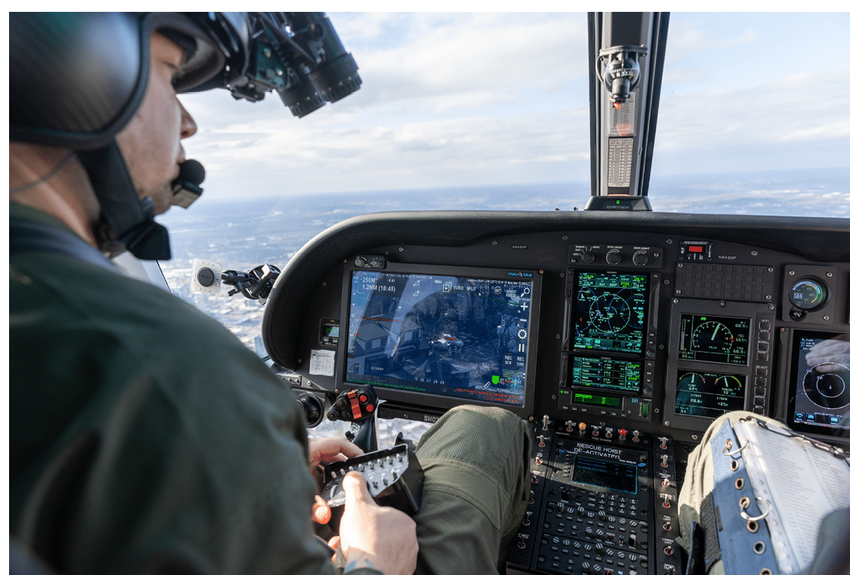 A person wearing a helmet and headset is operating the controls inside a helicopter cockpit with multiple digital displays showing flight information. Description generated by AI