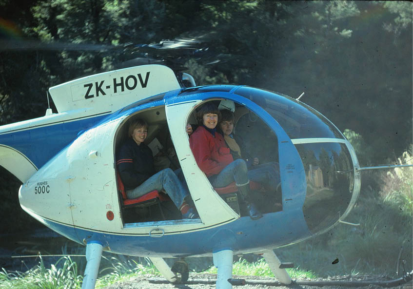 Three people sitting inside a small blue and white helicopter with the door open, smiling and looking at the camera. Description generated by AI