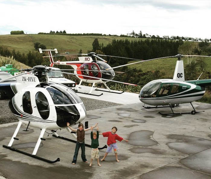 Three helicopters are parked on a concrete and gravel surface in a rural area with three children standing and waving in front of the nearest helicopter. Description generated by AI