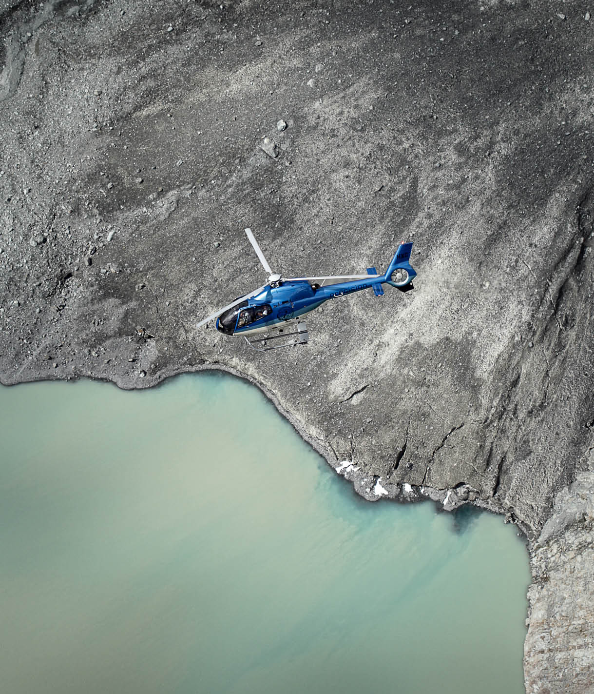 A blue helicopter is parked on a rocky, uneven surface with a mix of dark and light gray tones. Description generated by AI
