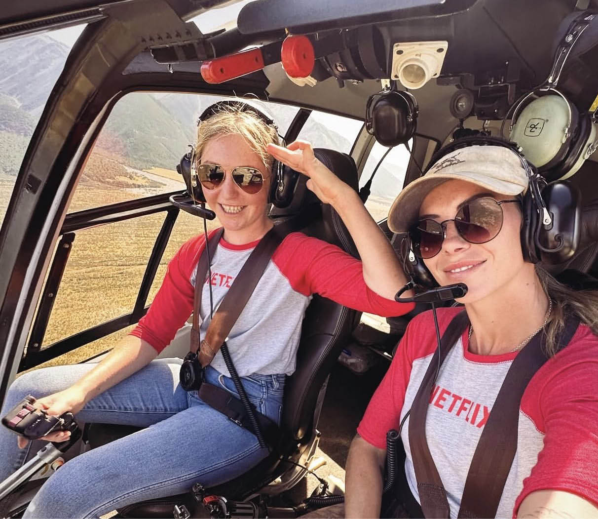 Two adults wearing matching red and gray Netflix shirts and sunglasses are smiling inside a helicopter cockpit, with one person holding the controls and the other taking a selfie. Description generated by AI