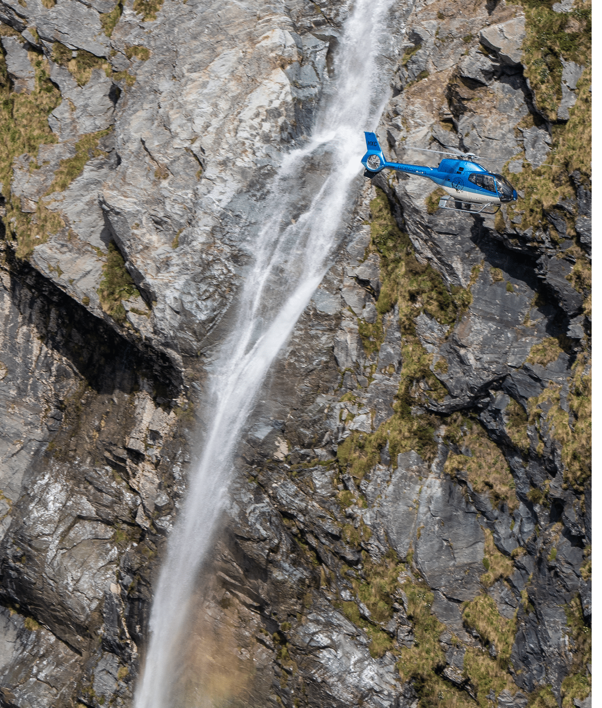 A blue helicopter is flying near a tall, narrow waterfall cascading down a rocky cliff. Description generated by AI