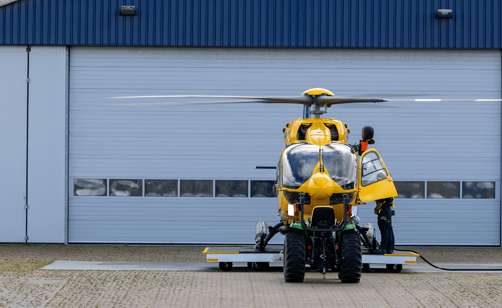 A yellow helicopter is parked on a platform in front of a large closed garage door while a person in dark clothing stands near the open side door of the helicopter. Description generated by AI