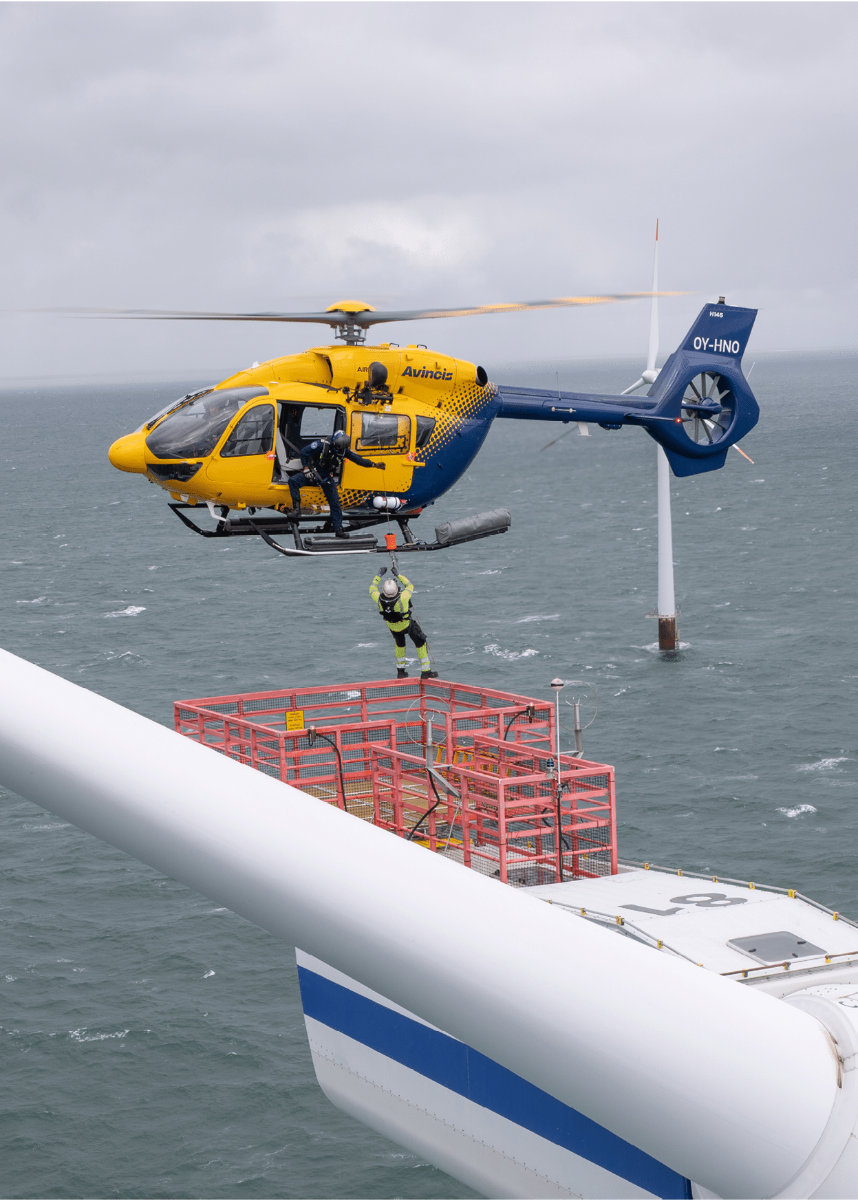 A helicopter hovers above the ocean near a wind turbine while a person in safety gear is being lowered onto a platform attached to the turbine. Description generated by AI