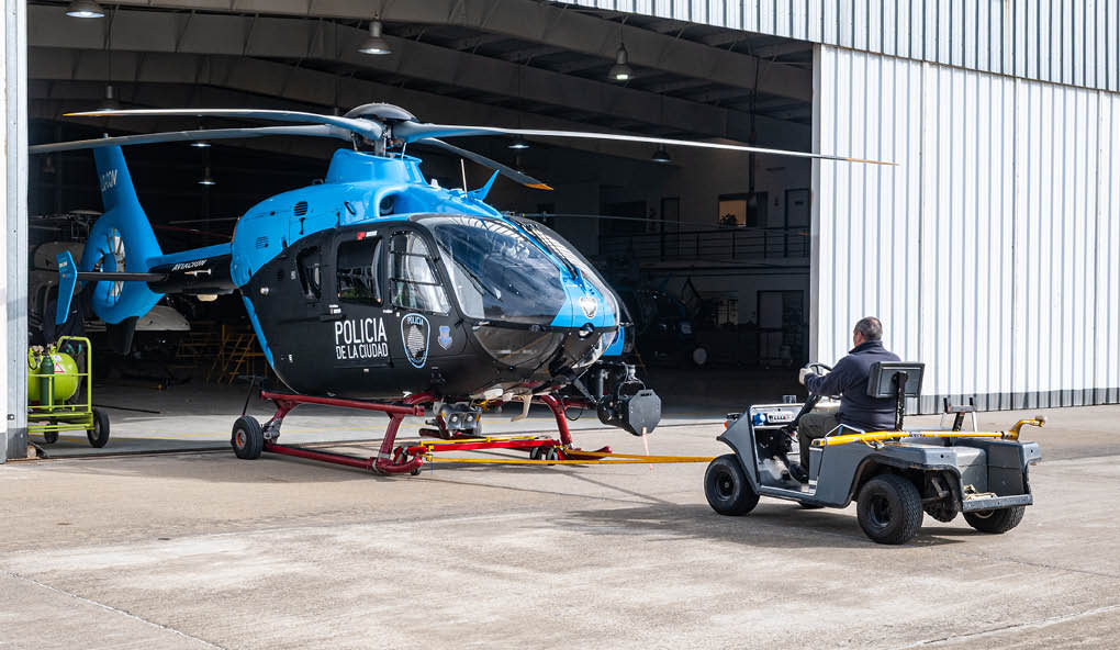 A blue and black helicopter is parked inside a large hangar with overhead lights visible. Description generated by AI