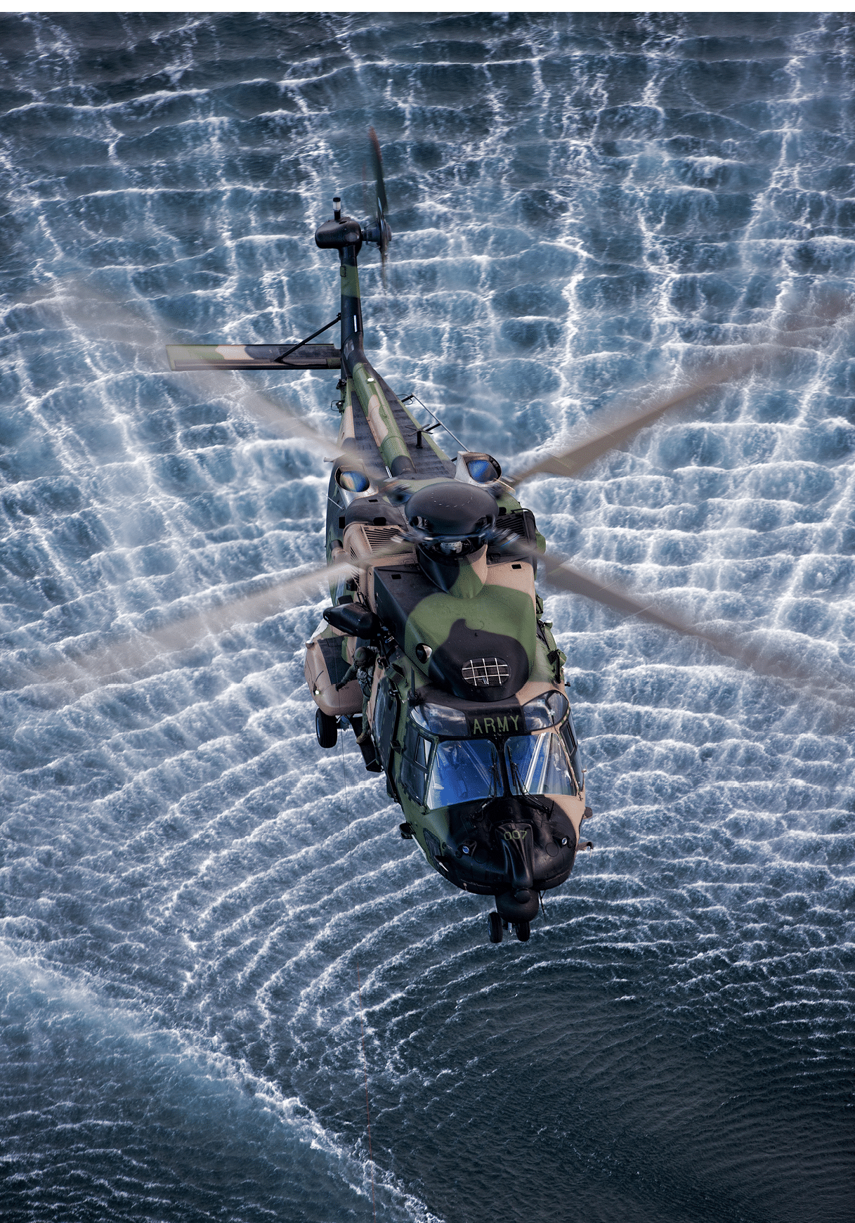 A military helicopter hovers above water, creating circular ripples with its spinning rotor blades. Description generated by AI