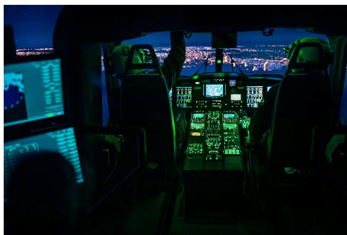 Two people are seated inside a dimly lit aircraft cockpit at night with illuminated control panels and city lights visible through the windows. Description generated by AI