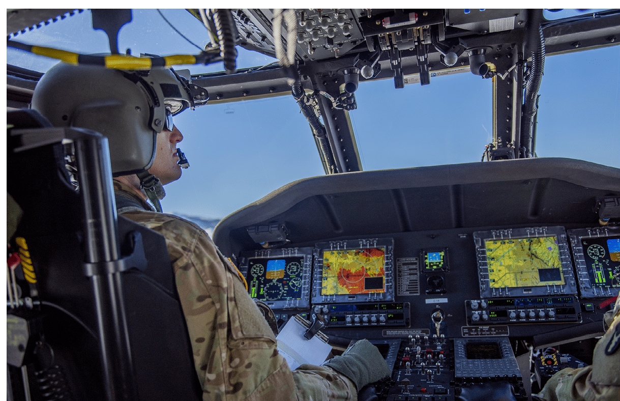 A person wearing a headset and camouflage clothing is seated in the cockpit of an aircraft, operating controls and monitoring multiple digital screens displaying flight and navigation information. Description generated by AI