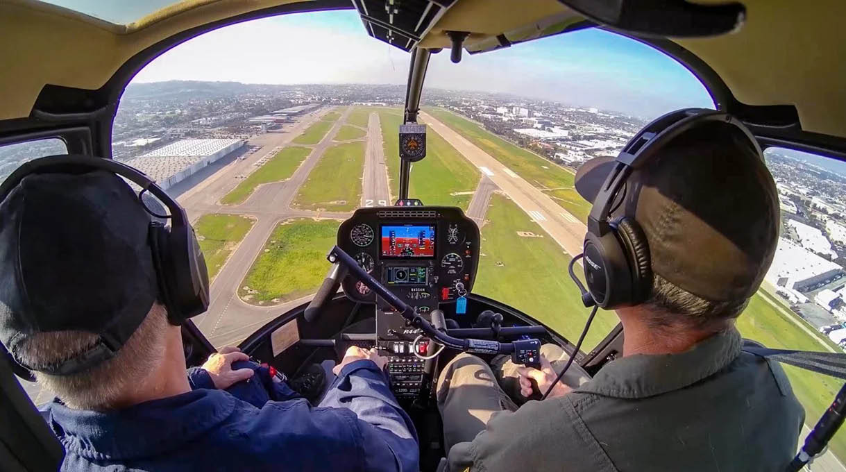 Two people wearing headsets are seated inside a small aircraft cockpit preparing to land on a runway visible through the windshield. Description generated by AI