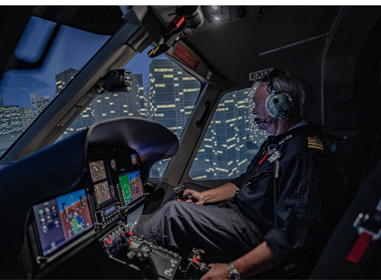 A person wearing a headset and dark clothing is seated in a cockpit at night, operating controls with illuminated city buildings visible through the windows. Description generated by AI