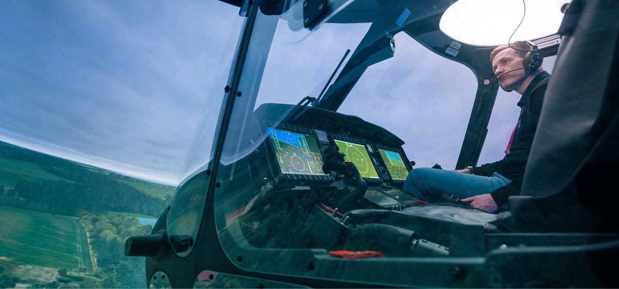 A person wearing a headset is seated inside a helicopter cockpit with visible control panels and a landscape below. Description generated by AI