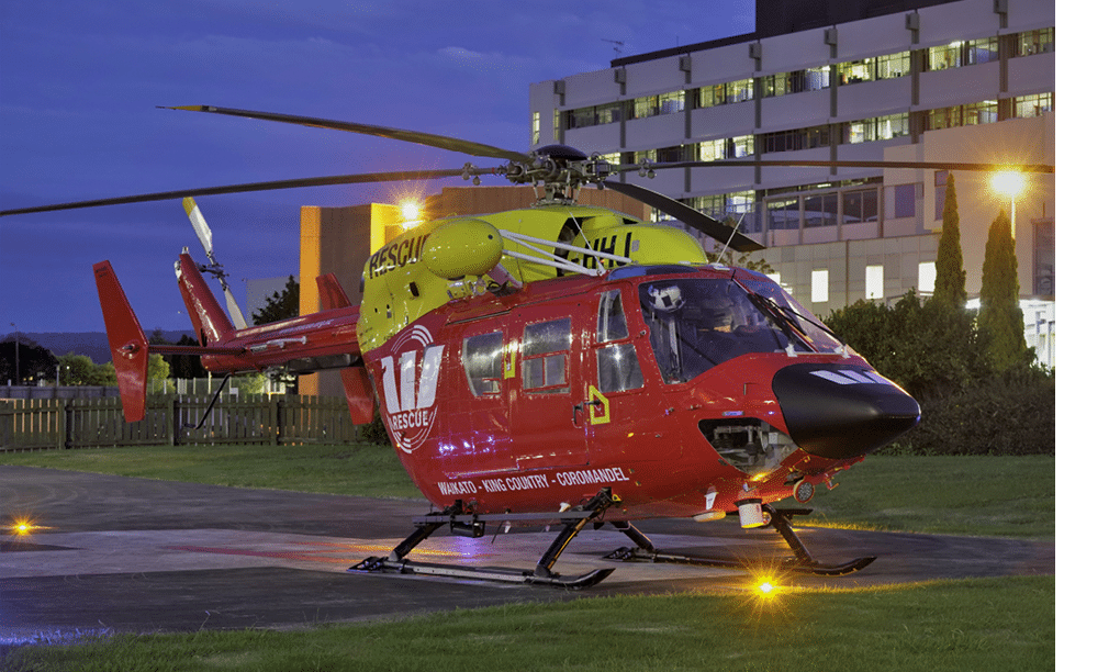 A rescue helicopter is parked outdoors near buildings under a darkening sky with its rotor blades visible. Description generated by AI