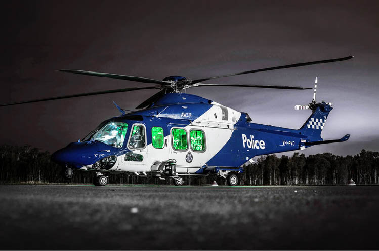 A blue and white police helicopter is shown on the ground with its rotor blades extended against a dark sky background. Description generated by AI