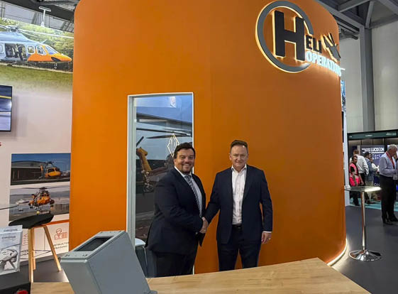 Two adults in suits are shaking hands and smiling in front of an orange wall with a partially visible logo and a small window showing a helicopter inside a modern indoor setting. Description generated by AI