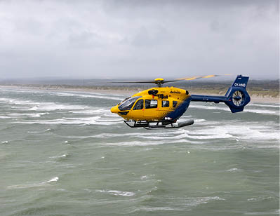 A yellow and blue helicopter is flying low over choppy ocean water near a sandy coastline under a cloudy sky. Description generated by AI