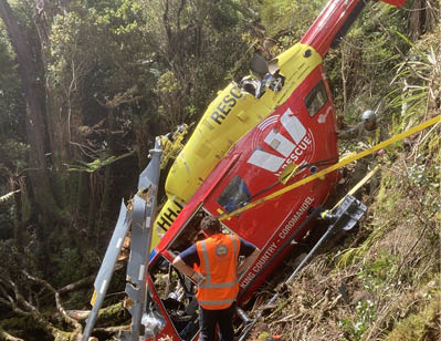 A person wearing an orange safety vest is standing next to a crashed red and yellow rescue helicopter in a forested area. Description generated by AI