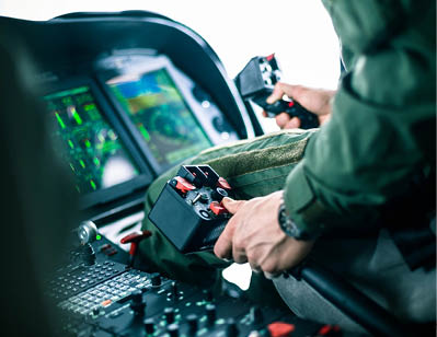 A person in a green uniform is operating controls inside a cockpit with multiple digital screens and buttons. Description generated by AI