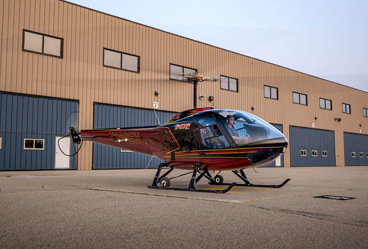 A small red and black helicopter is parked on a tarmac in front of a large industrial building with multiple windows and blue garage doors. Description generated by AI