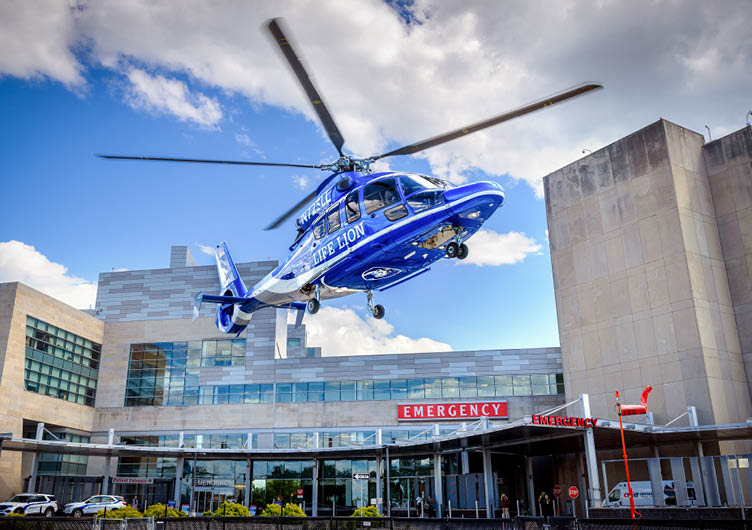 A blue and white medical helicopter is hovering above the emergency entrance of a modern hospital building under a partly cloudy sky. Description generated by AI