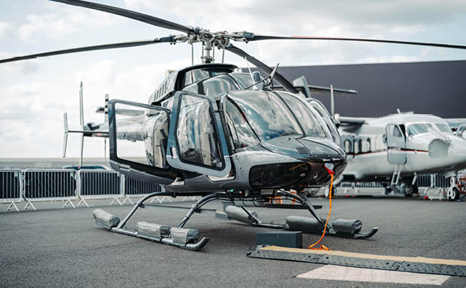 A sleek black helicopter is parked on a tarmac with a small airplane visible in the background under a cloudy sky. Description generated by AI