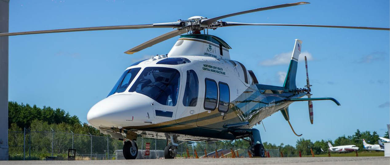 A white and teal helicopter is parked on a tarmac under a clear blue sky with trees and small aircraft in the background. Description generated by AI