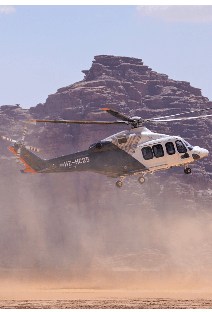 A helicopter is flying low over a dusty desert landscape with large rocky cliffs in the background. Description generated by AI