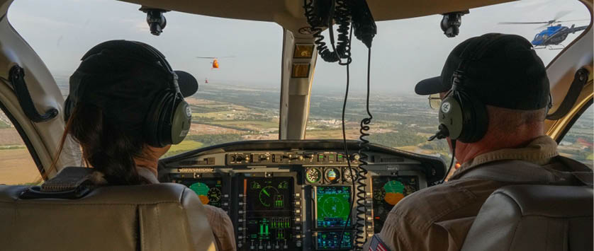 Two people wearing headsets and caps are seated in the cockpit of an aircraft with a view of the landscape through the windshield. Description generated by AI
