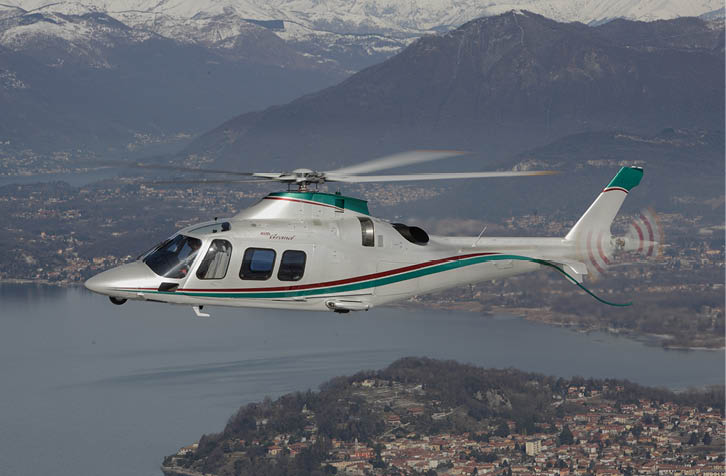 A white and green helicopter is flying over a lake with mountains and a town visible in the background. Description generated by AI