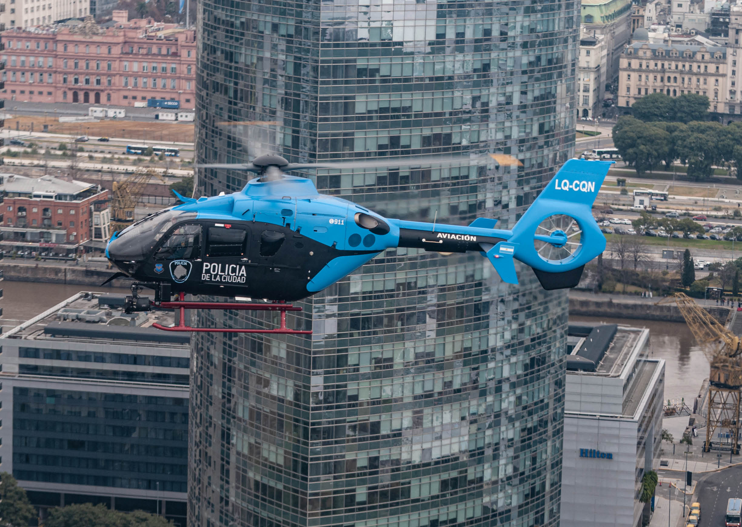 A blue and black police helicopter is flying in front of tall glass and brick buildings in an urban area. Description generated by AI