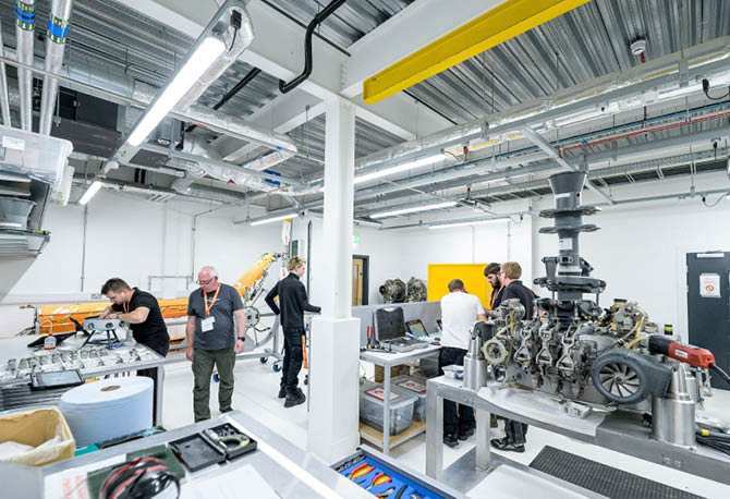 People in a bright industrial workshop are working on mechanical parts and engines with tools and computers on workbenches. Description generated by AI