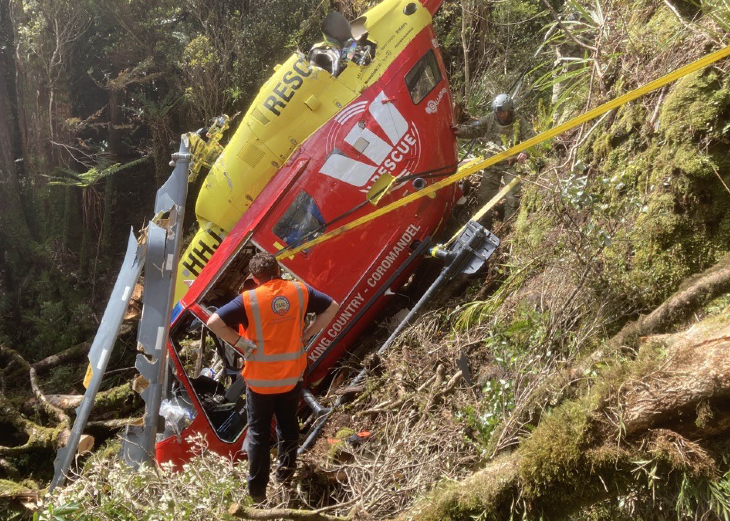 A rescue helicopter is tilted on its side on a steep, wooded hillside with people in safety vests and helmets attending to the scene. Description generated by AI