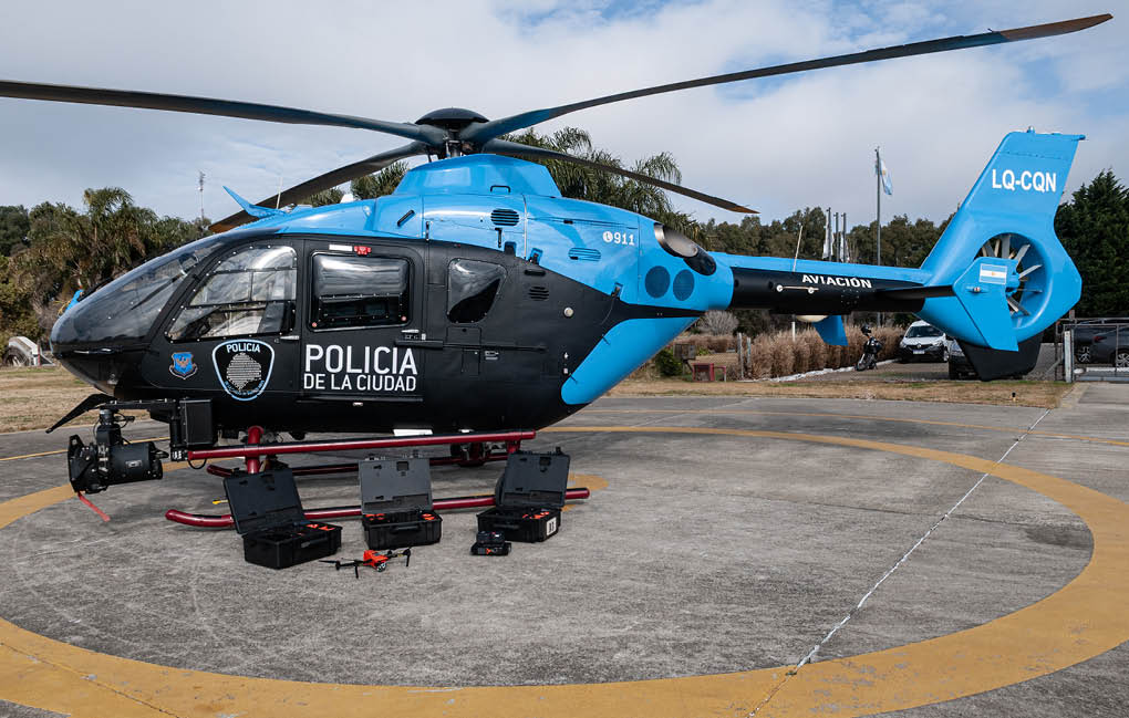 A blue and black police helicopter is parked on a helipad with several open black cases and a small drone placed on the ground nearby. Description generated by AI