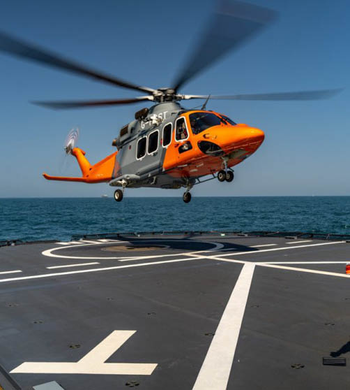 A person wearing a red helmet and green flight suit is guiding an orange and gray helicopter as it hovers above a ship's helipad on the ocean. Description generated by AI