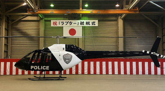 A black and white police helicopter is parked indoors in front of a red and white striped barrier with a Japanese flag and a banner with Japanese text hanging above. Description generated by AI