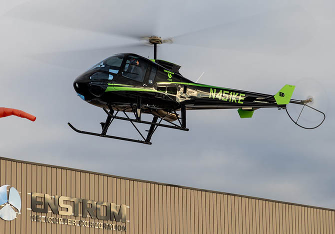 A black and green helicopter is hovering near a building with a windsock visible on the left side. Description generated by AI