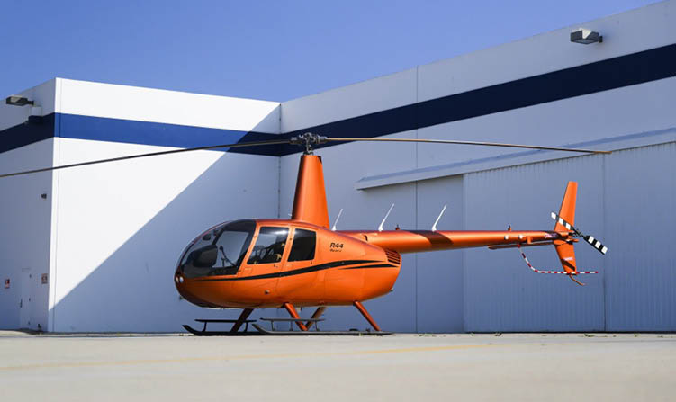 An orange helicopter is parked on a concrete surface next to a large white building under a clear blue sky. Description generated by AI