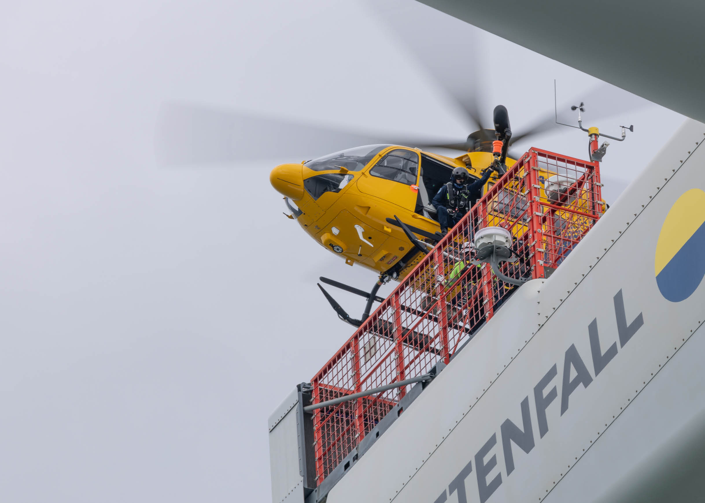 A yellow helicopter is hovering near a platform with red railings attached to a large structure labeled “VATTENFALL" where two people in safety gear are standing. Description generated by AI