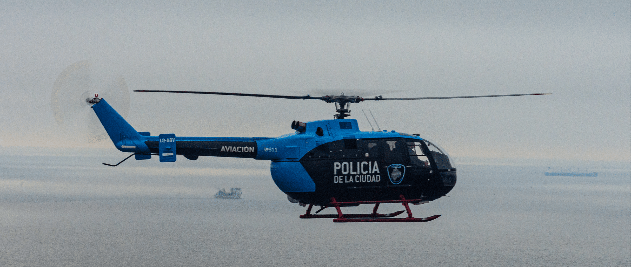 A blue and black police helicopter with red skids is flying over a body of water with ships visible in the distance. Description generated by AI