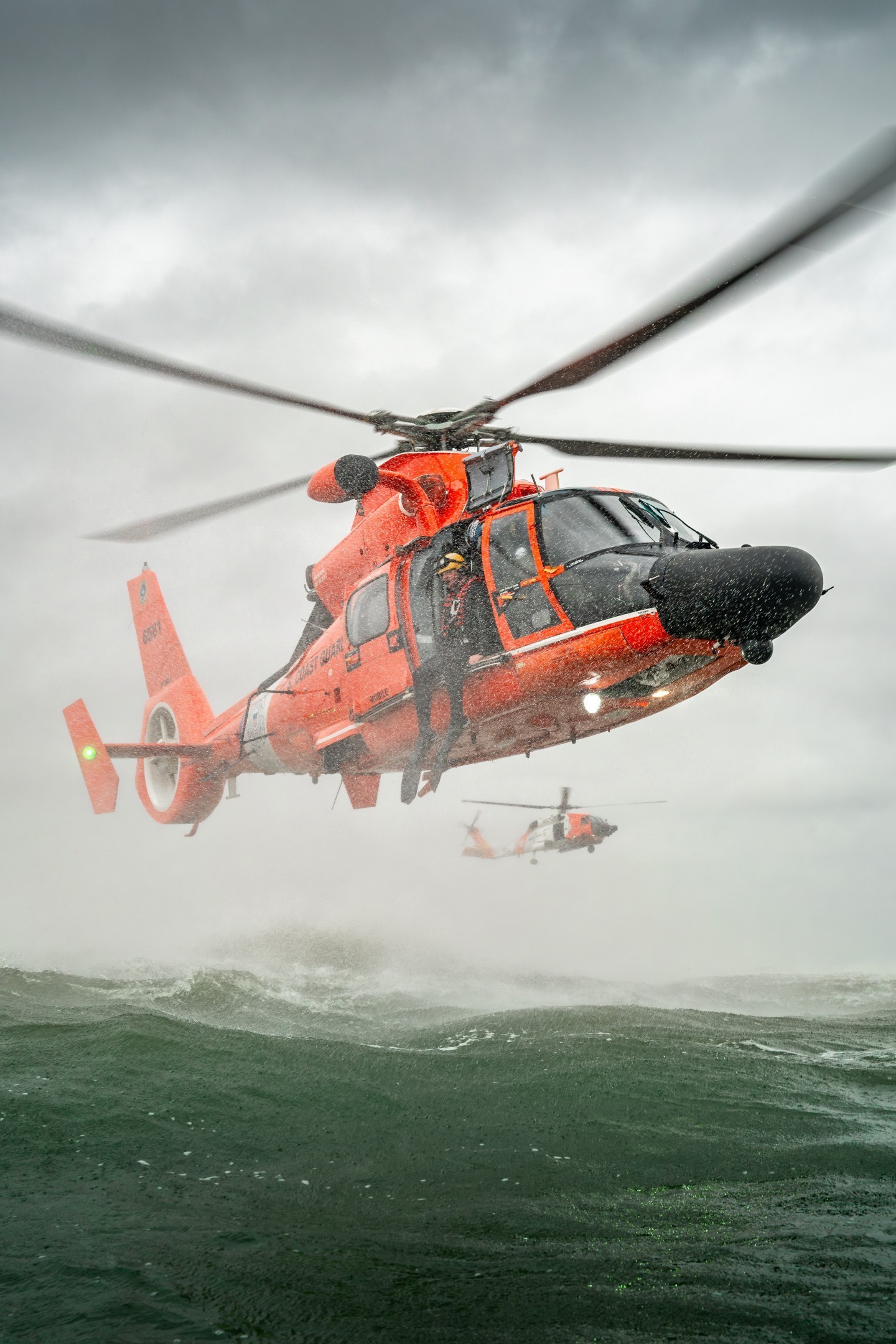 A Coast Guard Rescue Swimmer prepares for a Free Fall Deployment from a MH-65E during a training exercise while a MH-60T is looking from
afar.