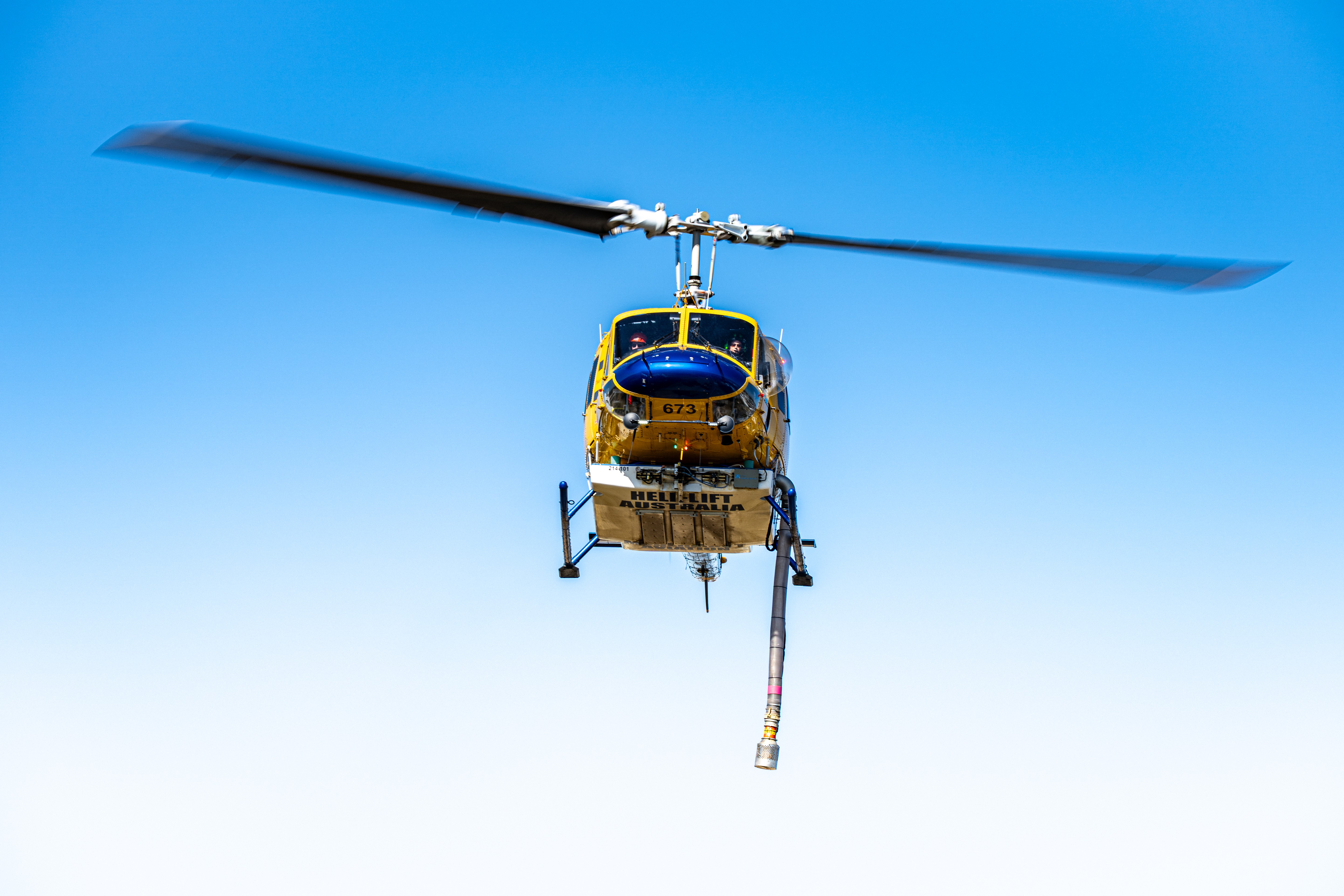 Bell 214 Helitack 673 arrives low and steady over Warradale Park, settling onto the grass for a brief but vital refuelling stop. The heavy helicopter’s broad rotor blades dominate the frame, frozen mid-turn as ground crew prepare to service the aircraft. Sunlight catches the fuselage, highlighting the work-worn paint and the unmistakable strength of a machine built for demanding conditions. Around it, the quiet suburban park contrasts sharply with the urgency of the mission, transformed into a temporary staging point for aerial firefighting. Every detail speaks of efficiency and readiness — the aircraft positioned with precision, the pause measured in minutes, not moments. Soon, fuel tanks will be full and 673 will lift again, returning to the fireground. The image captures a calm interval between intense operations, showing the essential logistics behind bushfire response and the power and presence of Helitack 673 at rest, but never idle.