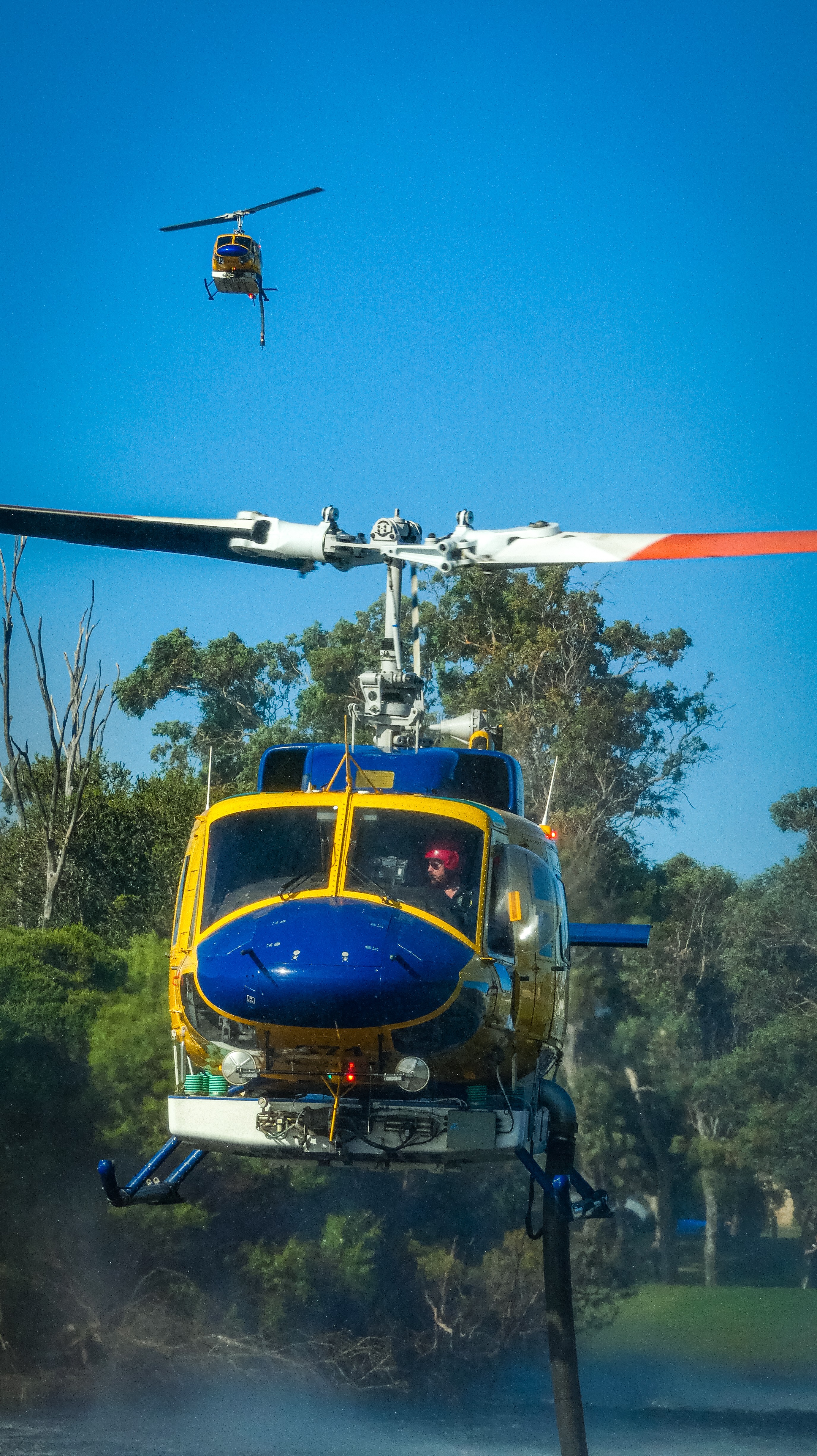 Bell 214 Helitack 674 hovers precisely over the water at Warradale Park, its powerful rotors flattening the surface into rippling rings of spray. The snorkel hangs steady beneath the aircraft, drawing water in seconds as the helicopter balances effortlessly just metres above the pond. Framed by trees and open parkland, the scene contrasts raw mechanical power with an otherwise quiet suburban setting. Rotor wash bends the surrounding grass and sends mist drifting across the water, capturing the intensity of aerial firefighting at close range. This brief moment is all business — no wasted movement, no hesitation — as 674 turns an everyday park into a vital supply point. Within moments the tank is full, and the helicopter will climb away toward the fireground. The image freezes that split second where skill, timing, and strength meet, highlighting the critical role of aerial crews and machines in protecting communities during bushfire operations.