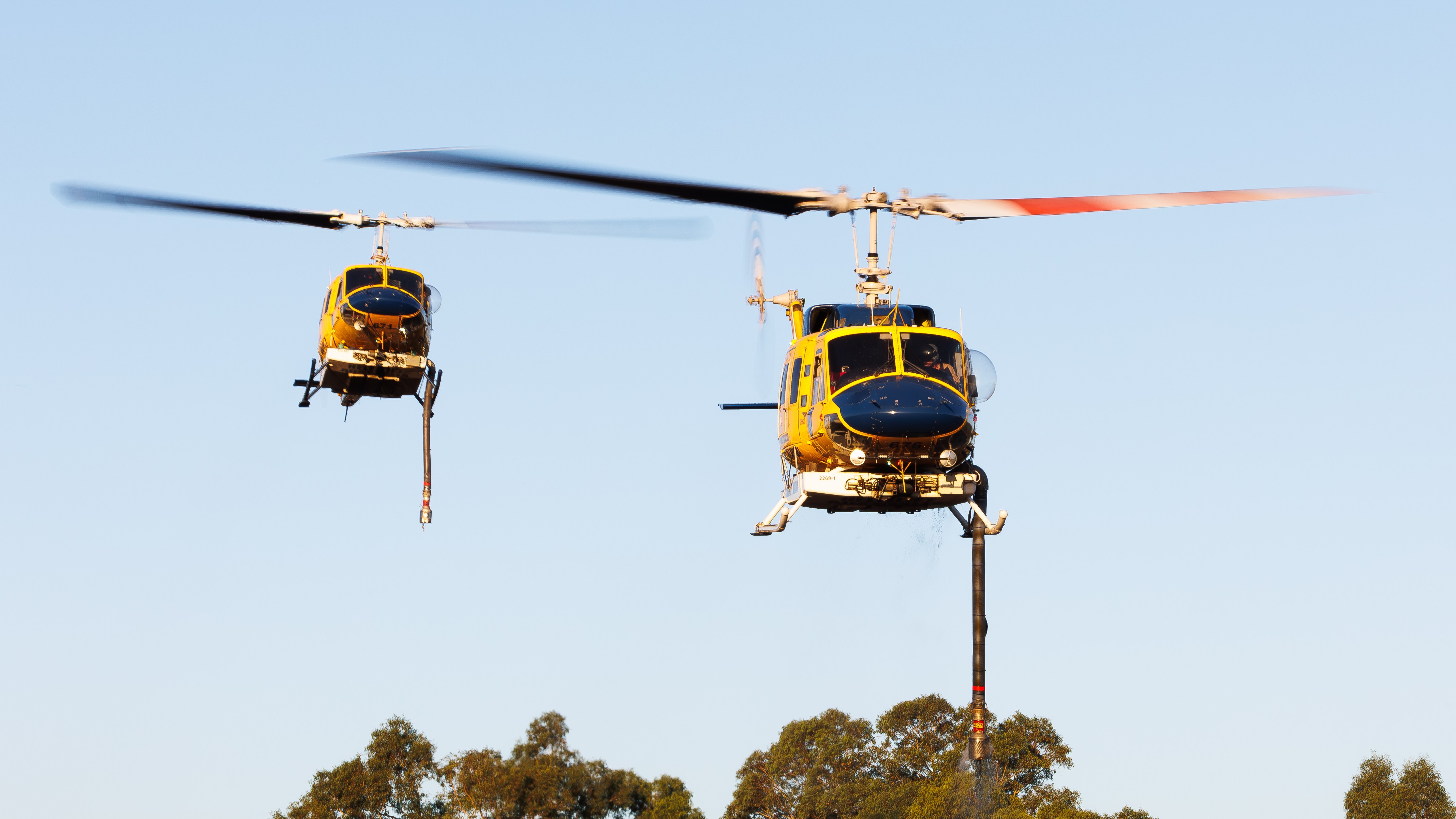 At a local dam, two Mcdermott Bell's 214B helicopters, HLTK676 and HLTL671, hover low over the water, working with speed and precision as an out-of-control bushfire burns nearby.