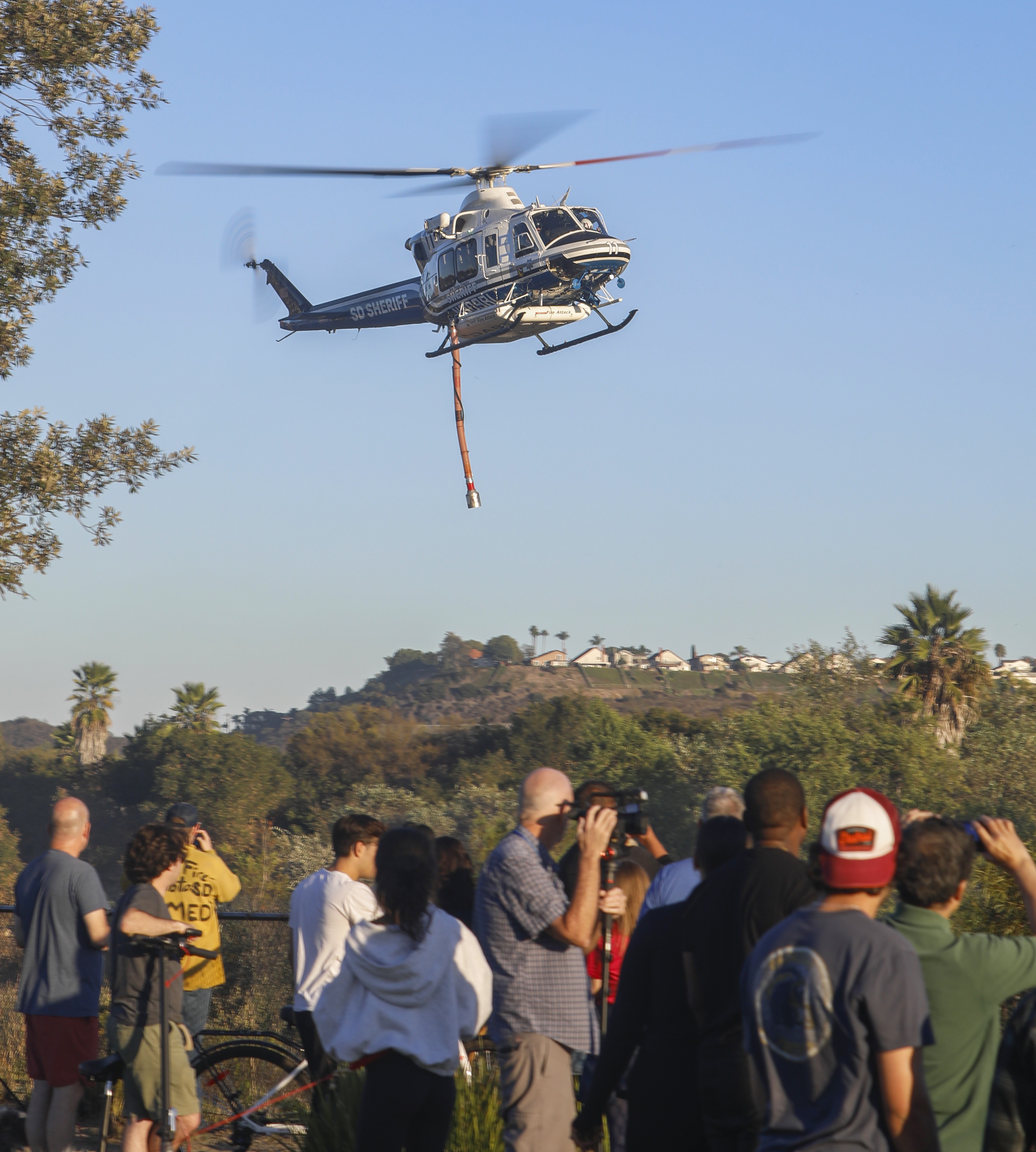 The public watches as Copter 11 from the San Diego County Sheriff's Dept. returns from a water dip during a small vegetation fire in the 4S Ranch area of San Diego, CA in October 2025.